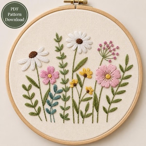 Könnte beinhalten: Ein runder Stickrahmen aus Holz mit cremefarbenem Stoff und einem handgestickten Blumenmuster. Die Stickerei zeigt weiße Gänseblümchen, rosa Kosmeen, kleine gelbe Blumen und grünes Blattwerk. Ein kreisförmiges Abzeichen zeigt den Text PDF Pattern Download.