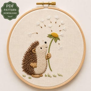 May include: A circular wooden embroidery hoop containing a fabric panel with a hand-stitched design of a brown hedgehog holding a yellow dandelion flower with white seeds blowing away. A green badge in the corner reads PDF PATTERN DOWNLOAD.