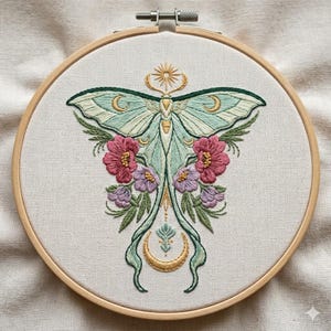 May include: Hand-stitched embroidery art in a wooden hoop. The design features a detailed moth with teal wings, adorned with gold accents and surrounded by pink and purple flowers. The background is a neutral fabric.
