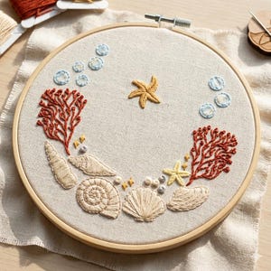 May include: A circular wooden embroidery hoop containing a beige fabric panel with hand-stitched ocean-themed designs. The embroidery features two red coral branches, various beige seashells, two yellow starfish, and light blue circular bubbles.