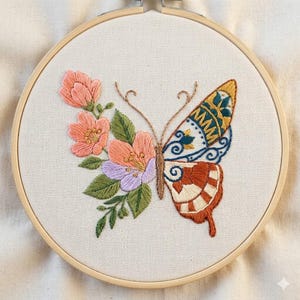 May include: A circular wooden embroidery hoop containing a fabric panel with hand-stitched needlework. The design features a butterfly with one wing composed of pink, peach, and lavender flowers with green leaves, and the other wing patterned.