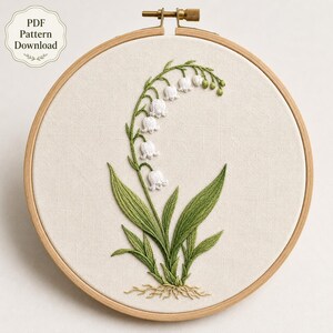 May include: A circular wooden embroidery hoop containing a piece of off-white fabric with a hand-stitched design of a lily of the valley plant. The plant features white bell-shaped flowers and green leaves. Text in the corner reads PDF Pattern Download.