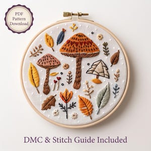Peut inclure: Un tambour à broder contenant un tissu blanc avec des motifs brodés de divers champignons, feuilles et petites fleurs dans des tons orange, marron, jaune et vert. Un badge indique PDF Pattern Download et le texte en bas indique DMC & Stitch Guide Included.