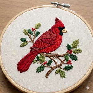 May include: Embroidered cardinal bird art in a wooden hoop. The red bird is perched on a brown branch with green leaves and red berries. The background is a light beige fabric. The embroidery is detailed and textured.