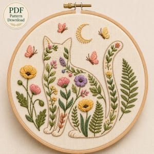 Peut inclure: Un tambour à broder contenant un panneau en tissu avec une silhouette de chat remplie de motifs floraux et botaniques colorés. Le design comprend des fleurs jaunes, roses et violettes, des feuilles vertes, des papillons et un croissant de lune.