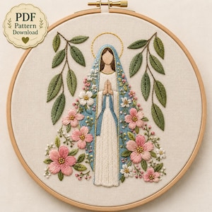 May include: A circular wooden embroidery hoop containing a fabric panel with a hand-stitched figure in a white gown and blue cloak, surrounded by pink flowers, white blossoms, and green leaves. A gold halo is stitched above the figure's head.
