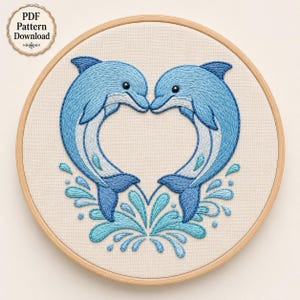 Op de afbeelding: Een borduurring met een stuk stof met een geborduurd ontwerp van twee blauwe dolfijnen die naar elkaar toe gericht zijn in een hartvorm, omringd door lichtblauwe waterspatten. Een rond label linksboven bevat de tekst PDF Pattern Download.
