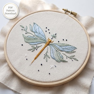 May include: A round wooden embroidery hoop containing off-white fabric with a hand-stitched dragonfly design. The dragonfly has a golden yellow body and light blue and green wings, surrounded by small black dots and white cross stitches. Text reads PDF Pattern Download.