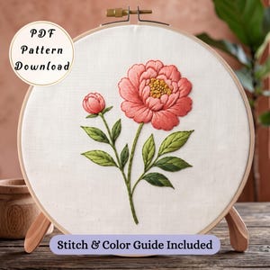 Puede incluir: Un bastidor de bordado redondo de madera con tela blanca que presenta una peonía de color rosa coral, un pequeño capullo y hojas verdes bordadas a mano. Una insignia circular dice PDF Pattern Download y un banner Stitch & Color Guide Included.