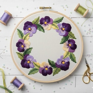 以下が含まれることがあります： 紫と黄色のパンジーと緑の葉が刺繍された花のリース。木製の刺繍枠に入っています。糸巻きや金色のハサミなどのクラフト用品が見えます。