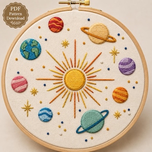 May include: A circular wooden embroidery hoop containing a fabric panel with a hand-stitched solar system design. The center features a bright yellow sun with radiating orange and yellow rays, surrounded by various planets and stars in colorful thread.