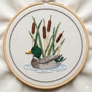 Op de afbeelding: Geborduurd kunstwerk met een wilde eend die in het water zwemt, omringd door lisdodden en riet. De eend heeft een groene kop, een bruin en grijs lichaam en een gele snavel. De borduursel zit in een houten hoepel met een neutrale stoffen achtergrond.