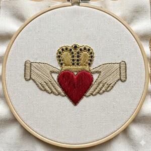 May include: Embroidered Claddagh design featuring a red heart, gold crown, and hands, set within a wooden embroidery hoop. The design is stitched on a cream-colored fabric, showcasing the traditional Irish symbol of love, loyalty, and friendship.