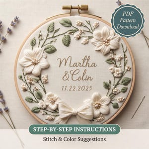 May include: A circular wooden embroidery hoop containing white fabric with a floral wreath design. The embroidery features white flowers, green leaves, and a central bow. The text reads Martha & Colin 11.22.2025. A needle rests nearby.