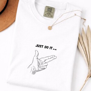 Op de afbeelding: Wit T-shirt met de tekst "JUST DO IT in ME" en een handgebaar illustratie. Een gouden ketting met een ronde hanger en een bruine hoed zijn ook zichtbaar. Het shirt heeft een label met "COMFORT COLORS".