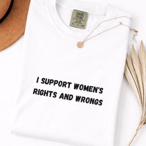 Op de afbeelding: Wit T-shirt met de zwarte tekst "I SUPPORT WOMEN'S RIGHTS AND WRONGS". Het shirt heeft een ronde hals en een label met de tekst "COMFORT COLORS". Een gouden ketting en een gedroogde plant zijn ook zichtbaar.