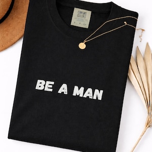 Op de afbeelding: Zwart T-shirt met de witte tekst "BE A MAN". Een gouden ketting met een ronde hanger ligt op het shirt. Een bruine hoed en een gedroogde plant staan op de achtergrond.