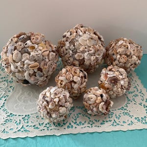 May include: A collection of seven decorative spheres, each meticulously covered in a variety of seashells. The shells are primarily neutral tones of beige, cream, and brown, creating a textured, natural aesthetic. The spheres vary in size, offering a unique coastal decor element.