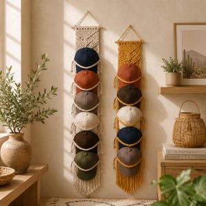 May include: Two vertical macrame wall organizers holding multiple baseball caps. The left organizer is cream-colored with caps in navy, rust, gray, white, brown, and olive. The right organizer is mustard yellow with caps in rust, brown, white, navy, and gray.