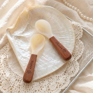 Puede incluir: Dos cucharas pequeñas con cuencos de concha iridiscente de color blanco roto y mangos de madera marrón lisa. El mango de una cuchara está grabado con el nombre Oliver y un icono de cupcake, mientras que la otra tiene el nombre Sophia y un molinillo.
