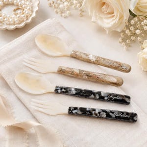 May include: Two spoons and two forks with cream-colored heads and patterned handles. The top two handles feature a brown and white marbled design with the names Isabella and Charlotte. The bottom two handles have a black and white marbled design with the names Sophia and Madeline engraved in white script.