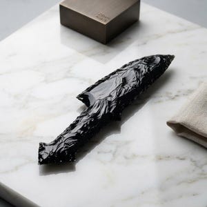 May include: A black obsidian arrowhead with detailed carvings is displayed on a white marble surface. A small, dark brown box with the text "Zidan Agate" is in the background, along with a folded beige cloth. The arrowhead is a fine example of craftsmanship.