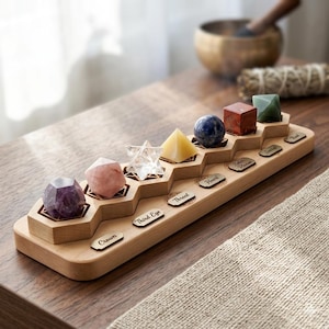 Può includere: Un espositore in legno con sette cristalli colorati di varie forme. I cristalli sono disposti su una piattaforma a gradini, con etichette che indicano i chakra. L'espositore è in legno chiaro e si trova su una superficie di legno.