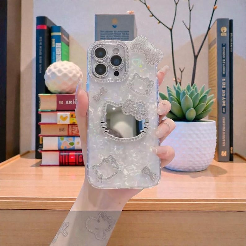 May include: A clear phone case with a pearlescent finish, decorated with sparkling rhinestones. The case features a Hello Kitty design, including a bow and the character's face. The case is designed to fit a smartphone.