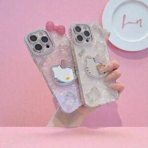 May include: Two decorative phone cases featuring a Hello Kitty design. One is pink with a bow, the other white with a bow. Both cases have a mirrored Hello Kitty face and are embellished with rhinestones.