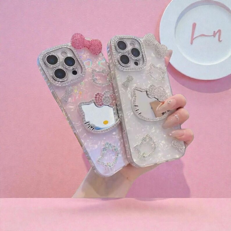 May include: Two decorative phone cases featuring a Hello Kitty design. One is pink with a bow, the other white with a bow. Both cases have a mirrored Hello Kitty face and are embellished with rhinestones.