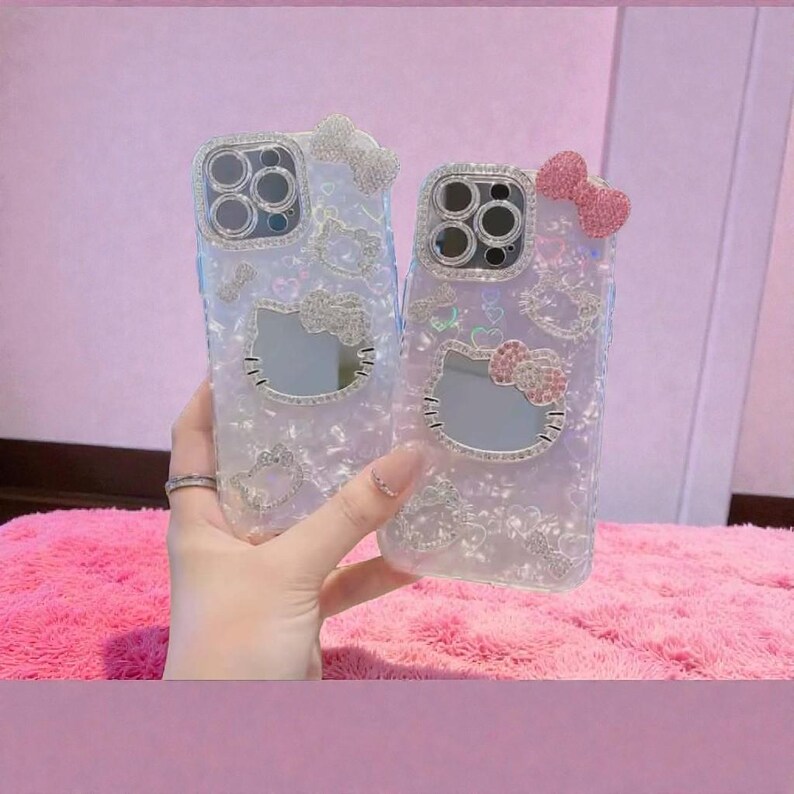 May include: Two clear phone cases with Hello Kitty designs. Each case features a bow at the top and a mirrored Hello Kitty face. The cases are embellished with rhinestones and glitter, creating a sparkling effect.