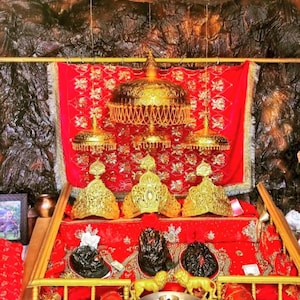 Puede incluir: Un santuario religioso ornamentado con detalles dorados y un fondo rojo. El santuario presenta coronas doradas, paraguas decorativos y estatuas. El fondo es una superficie oscura y texturizada, creando un contraste con los colores brillantes del santuario.