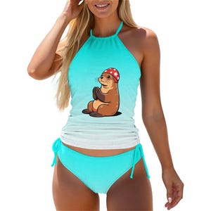 May include: A turquoise halter-neck swimsuit featuring a cartoon sea otter. The sea otter is brown and wears a red and white star-patterned hat. The swimsuit has a matching turquoise bottom with adjustable side ties.