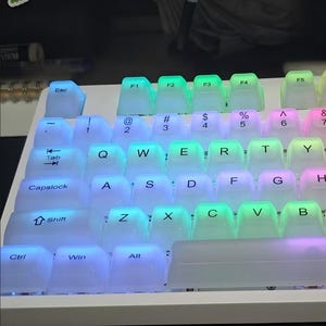 May include: A white mechanical keyboard with transparent keycaps illuminated by a rainbow of LED lights. The keys are arranged in a standard QWERTY layout, with function keys and modifier keys visible. The lighting creates a vibrant and colorful effect.