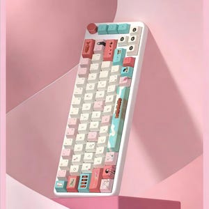 K&ouml;nnte beinhalten: Eine wei&szlig;e mechanische Tastatur mit pastellfarbenen rosa, blauen und wei&szlig;en Tastenkappen. Die Tastatur hat ein einzigartiges Design mit individuellen Tastenkappen mit Illustrationen. Die Tastatur ist abgewinkelt und vor einem rosa Hintergrund platziert.