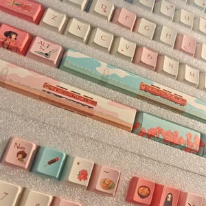 K&ouml;nnte beinhalten: Eine Sammlung von individuellen Tastaturkappen mit japanischen Motiven. Die Tastenkappen sind in Wei&szlig;, Rosa und Blau gehalten und zeigen Illustrationen von Landschaften, Essen und Charakteren. Die Tastenkappen haben japanischen Text und Symbole.