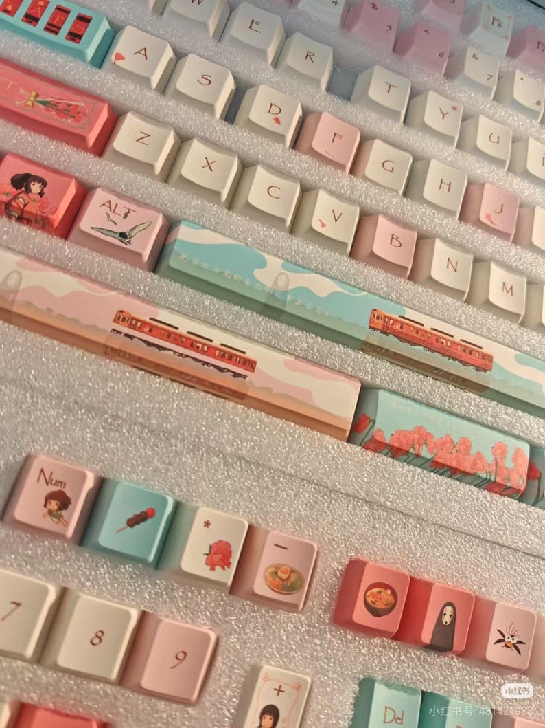 K&ouml;nnte beinhalten: Eine Sammlung von individuellen Tastaturkappen mit japanischen Motiven. Die Tastenkappen sind in Wei&szlig;, Rosa und Blau gehalten und zeigen Illustrationen von Landschaften, Essen und Charakteren. Die Tastenkappen haben japanischen Text und Symbole.