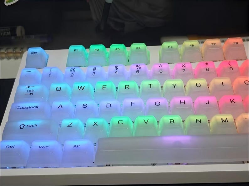 May include: A white mechanical keyboard with transparent keycaps illuminated by a rainbow of LED lights. The keys are arranged in a standard QWERTY layout, with function keys and modifier keys visible. The lighting creates a vibrant and colorful effect.