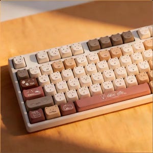 May include: A white mechanical keyboard with keycaps featuring cat faces in various shades of brown and beige. The space bar is a dusty rose color and has the text "Coffee Daily". The keyboard has a minimalist design.