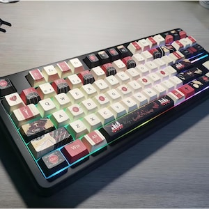 Può includere: Tastiera meccanica nera con tasti personalizzati. I tasti presentano un mix di colori bianco, rosso e nero con vari disegni. La tastiera ha una cornice nera con una striscia luminosa a LED colorata lungo il bordo inferiore. È visibile un estrattore di tasti.