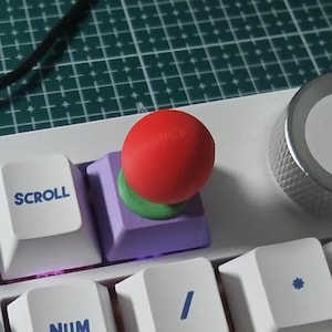 Puede incluir: Primer plano de un teclado mecánico blanco con teclas personalizadas. Una tecla roja esférica se asienta sobre una tecla púrpura, con una base verde. El teclado tiene teclas etiquetadas como "SCROLL" y "NUM". Se ve un dial plateado.