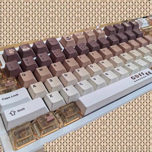 May include: A mechanical keyboard with a white frame and a gradient of brown keycaps. The keycaps transition from light beige to dark brown. Some keys have transparent accents. The space bar reads "COFFEE".