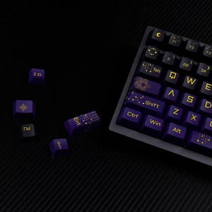 Könnte beinhalten: Nahaufnahme einer mechanischen Tastatur mit lila Tastenkappen und goldfarbenen Buchstaben. Mehrere Tastenkappen liegen um die Tastatur herum verstreut. Die Tastenkappen haben Himmelsmotive und Symbole. Die Tastatur befindet sich auf einer dunklen Oberfläche.