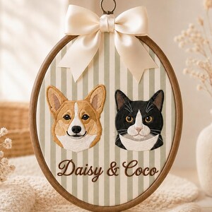 May include: An oval embroidery hoop featuring a light green and white striped fabric background. It displays embroidered portraits of a tan and white Corgi and a black and white cat, with the brown embroidered text Daisy & Coco below them.
