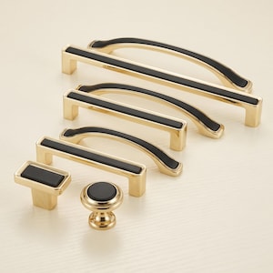 Puede incluir: Una colección de herrajes para muebles que incluye varios tiradores y pomos de metal en tono dorado. Cada pieza presenta una incrustación negra mate, rectangular o circular, centrada sobre la superficie dorada pulida y brillante.