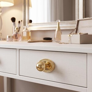 May include: A polished gold-toned spherical drawer knob mounted on an oval backplate with a textured, cross-hatched pattern. The knob is attached to a white wooden drawer front, featuring a smooth, rounded handle design with a reflective finish.