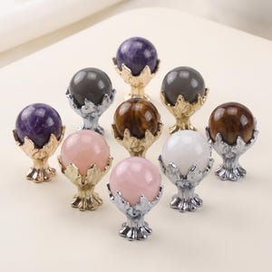 May include: A collection of nine spherical gemstone knobs, each set in a decorative metal base. The stones include purple amethyst, pink rose quartz, brown tiger's eye, and grey spheres. The metal bases feature a leaf-like design in gold or silver.