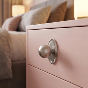 May include: A brushed silver spherical knob mounted on a vertical oval backplate with a textured beaded edge. The hardware is attached to a light pink wooden drawer front, featuring a smooth finish and a horizontal line across the center.