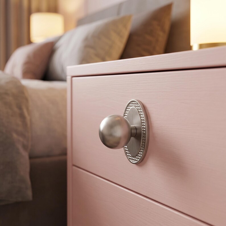 May include: A brushed silver spherical knob mounted on a vertical oval backplate with a textured beaded edge. The hardware is attached to a light pink wooden drawer front, featuring a smooth finish and a horizontal line across the center.