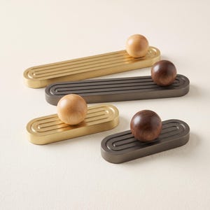 May include: Four cabinet pulls featuring elongated rectangular metal bases with rounded ends and grooved detailing. Two bases are finished in brushed gold and two in dark bronze, each topped with a smooth, spherical wooden knob.